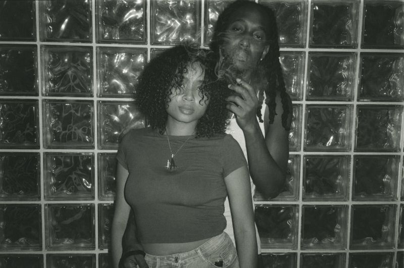 Black and white photo of an adult couple posing in front of a glass block wall; one is smoking with smoke visible in frame.