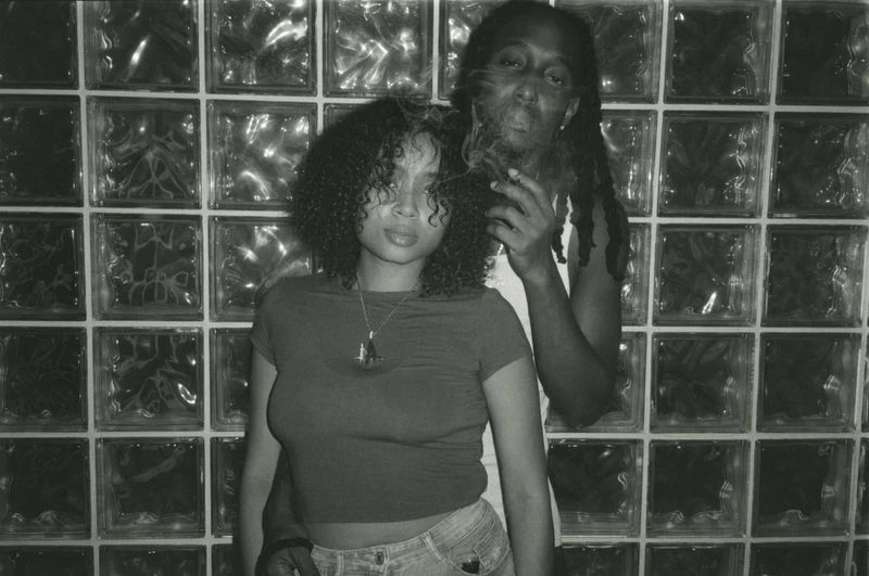 Black and white photo of an adult couple posing in front of a glass block wall; one is smoking with smoke visible in frame.