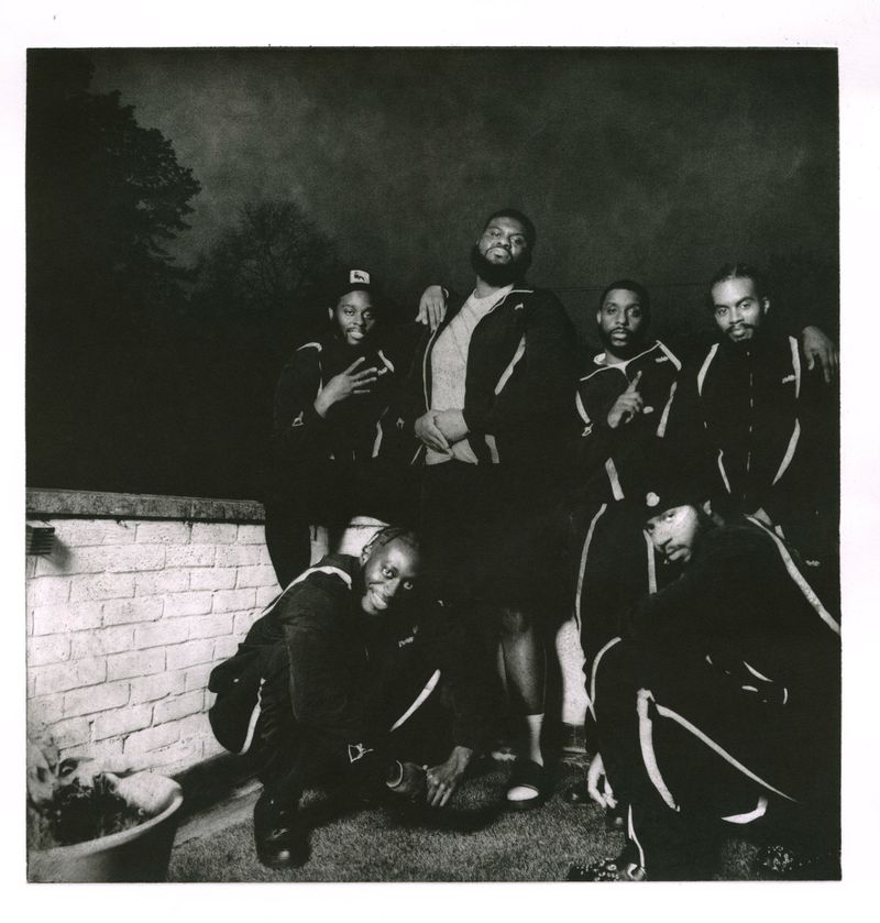 Monochrome group portrait of six African-descent men in tracksuits posing outside in dim lighting with a brick wall backdrop.