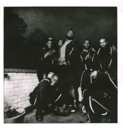 Black and white portrait of a group of Black men posing together outdoors at night wearing dark matching tracksuits.