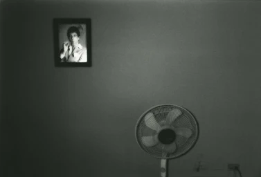 A framed portrait on a dimly lit wall positioned above a white electric standing fan with wall sockets visible below.