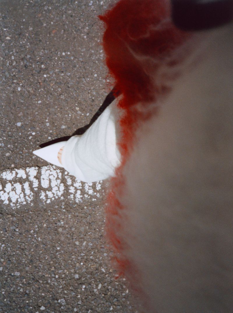 Aerial view of woman's legs and feet wearing white heels, standing on a gray concrete ground. Red and white fluffy material visible.