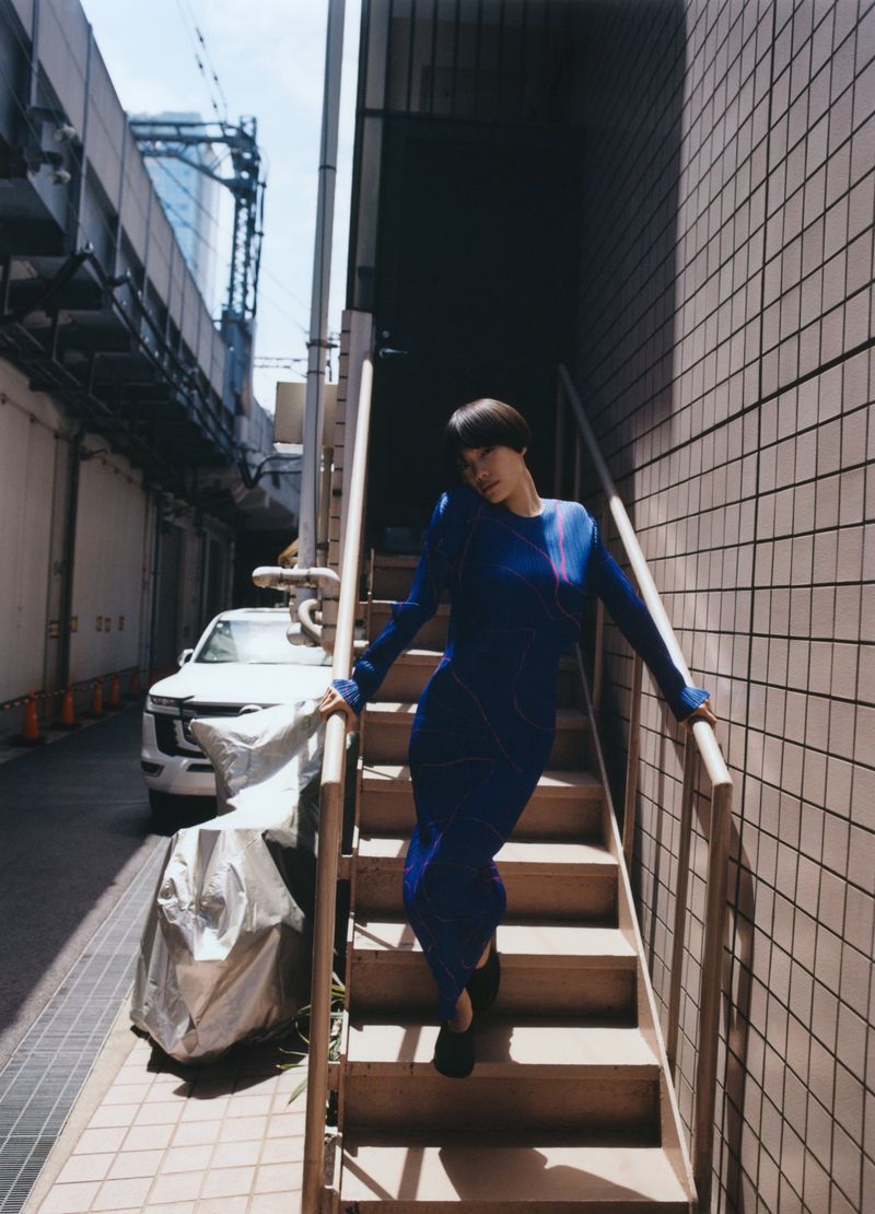 Woman in a blue dress descends outdoor steps in an urban alley with a white car and covered motorcycle visible in the background.