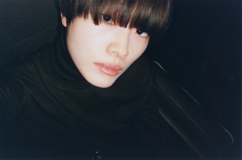 Close-up portrait of a woman with short brown hair, fair skin, and a black turtleneck against a dark background in low lighting.