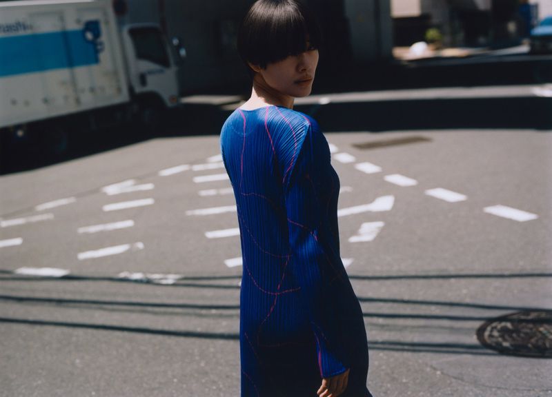 Adult female model wearing a blue pleated dress with pink details posing on an urban street with a truck in the background.