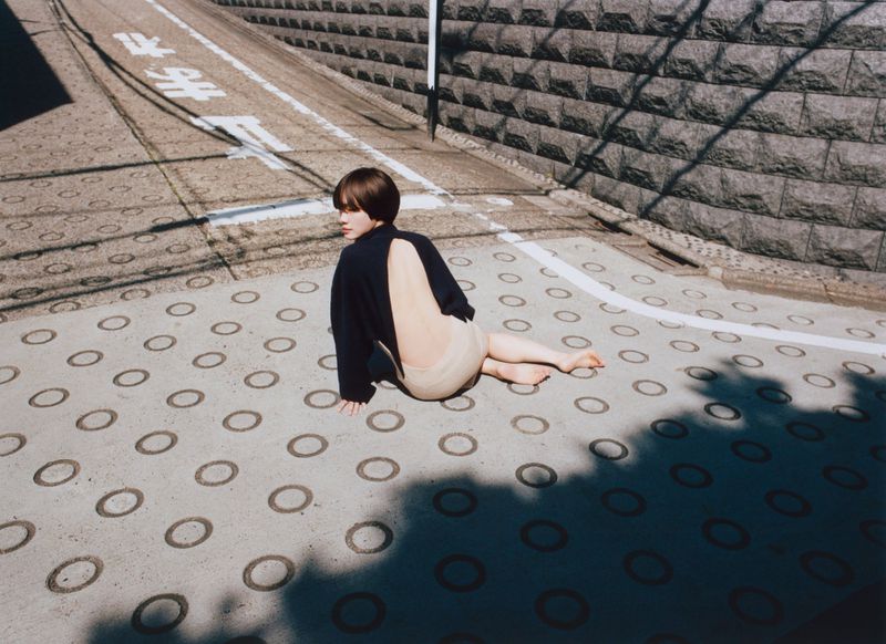 A woman with short brown hair sits on a concrete surface with ring patterns wearing a black sweater with an open back.