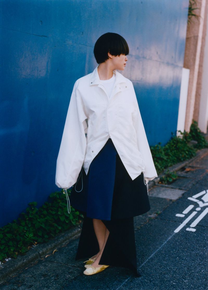 Woman in oversized white jacket and blue skirt against a blue wall. Fashion shot with an urban background, natural lighting.