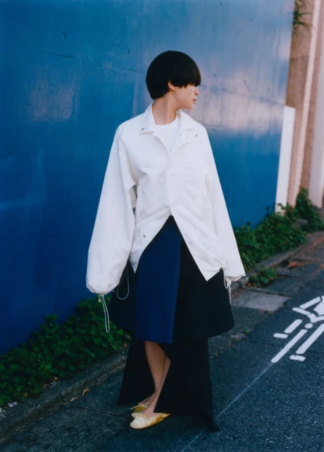 Woman in oversized white jacket and blue skirt against a blue wall. Fashion shot with an urban background, natural lighting.