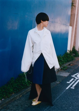 Woman in oversized white jacket and blue skirt against a blue wall. Fashion shot with an urban background, natural lighting.