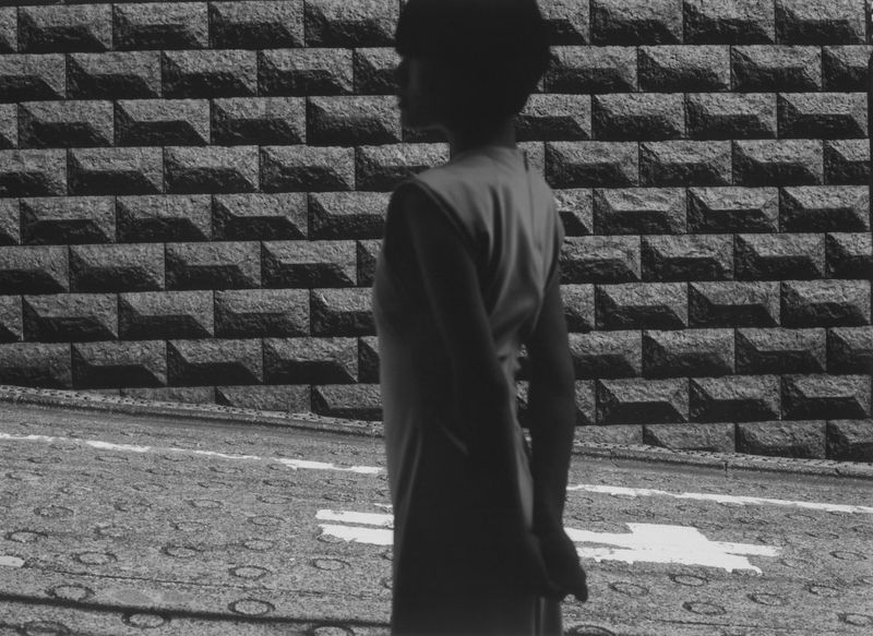Monochrome portrait of a solitary young person standing near a textured stone brick wall and patterned ground, captured in profile.