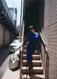 Woman with a short dark bob wearing a long cobalt blue dress standing on an outdoor staircase in an alley.