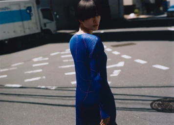 Model with short black hair standing in a street wearing a pleated royal blue long-sleeve dress with pink linear detailing.
