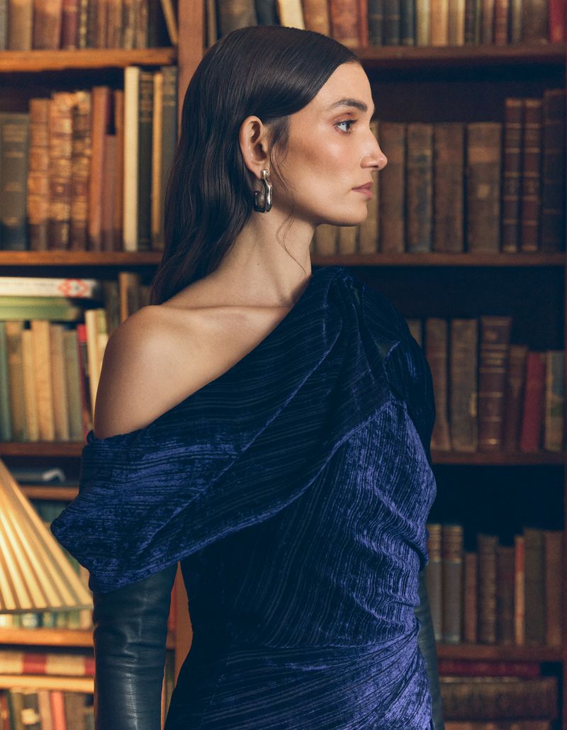 A woman with dark hair and a navy velvet dress with books in the background poses in a studio portrait with medium framing and eye-level angle.