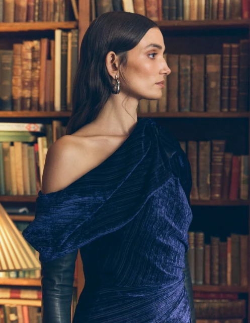 A woman with dark hair and a navy velvet dress with books in the background poses in a studio portrait with medium framing and eye-level angle.