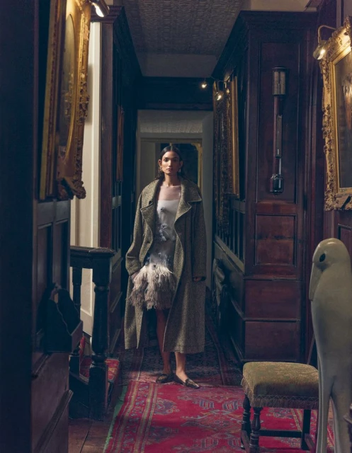 Elegant woman in coat and dress poses in an opulent hallway with wood panelling, carpet, paintings, and a decorative ceramic bird.