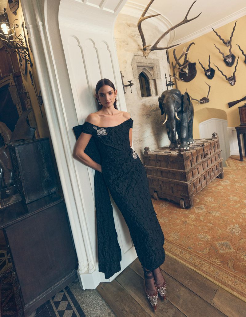 Woman in elegant black gown poses in a room with mounted antlers and a vintage elephant statue, highlighting a blend of fashion and decor.