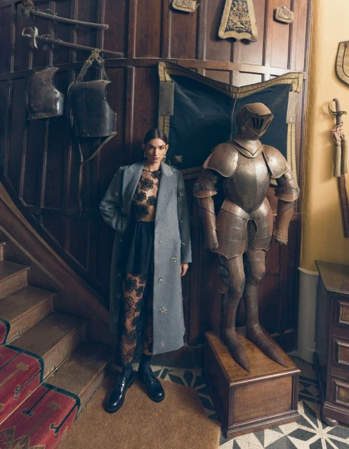 Woman poses in elegant floral top, black tights, and grey coat next to a knight's armor in a wood-paneled interior with carpeted stairs.