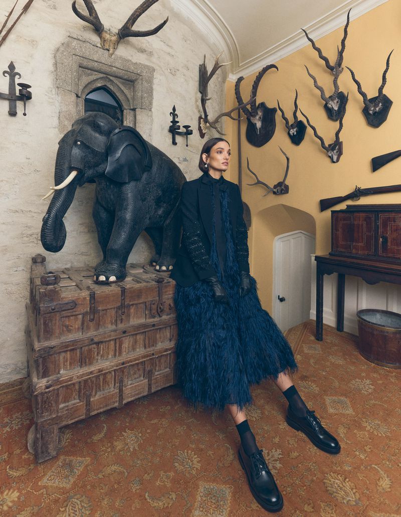 A fashionable woman in a blue dress sits next to an elephant statue in a stately room with yellow walls and mounted animal heads.