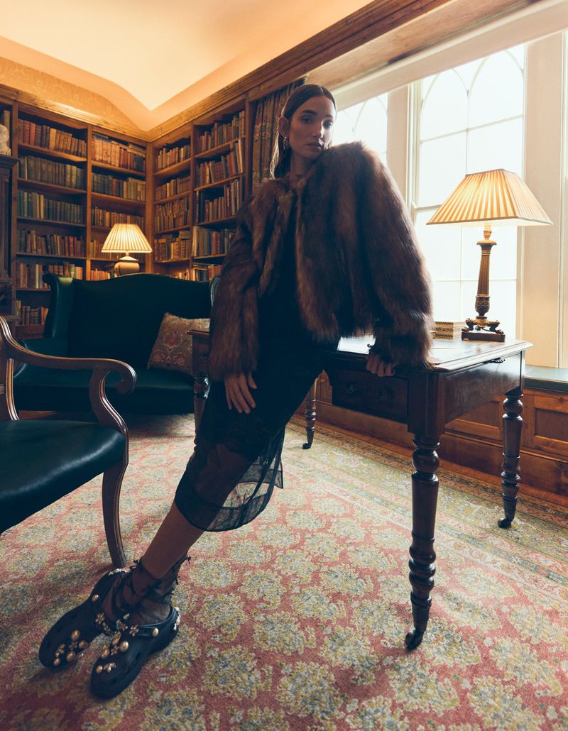 A woman in a fur coat and black dress leans on a wooden desk in a room with a large bookcase, creating a sophisticated fashion portrait.