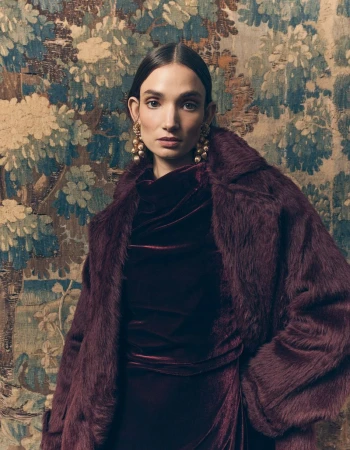 Model wearing a deep burgundy fur coat and velvet dress with statement earrings against an ornate tapestry backdrop.