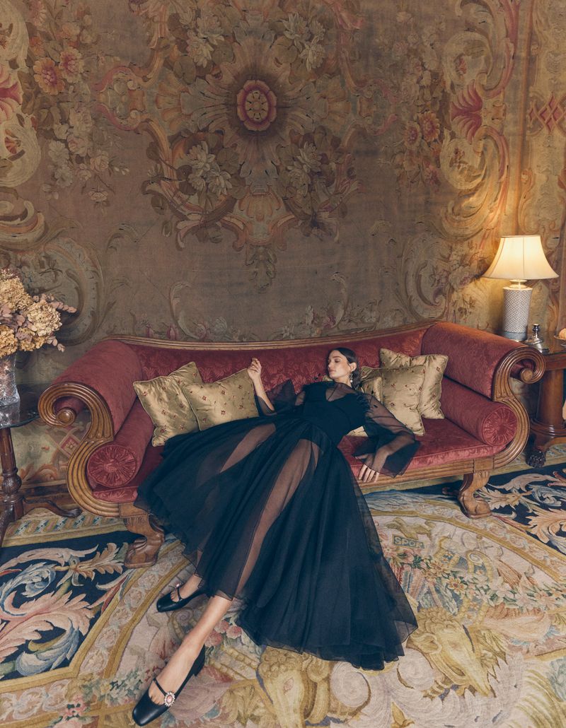 Elegant woman in black tulle dress poses on a red velvet couch against a floral tapestry background in a fashion photography setting.