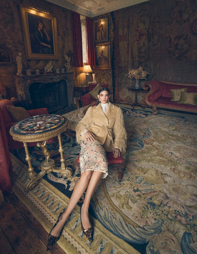 Indoor fashion portrait of a woman in a beige jacket and beaded skirt sitting on a red stool in a historic room.