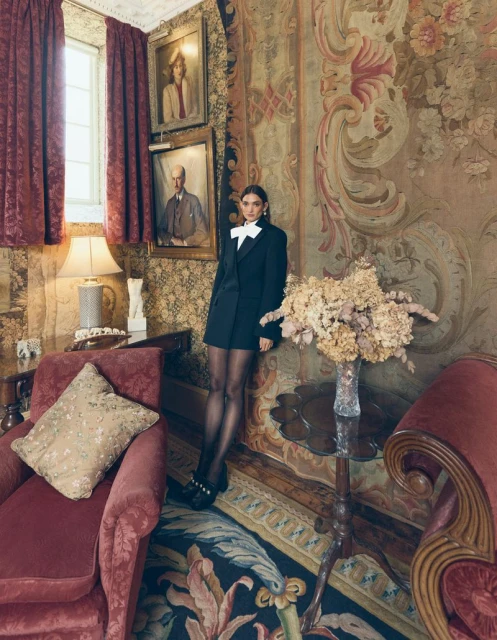 A woman in a black blazer dress poses in an ornate room with paintings, a table with flowers, and antique furniture, creating a vintage aesthetic.