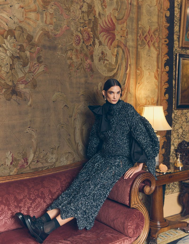 Woman in dark sweater dress and black shoes sits on a red velvet couch in a room with a tapestry and dark wall decor.