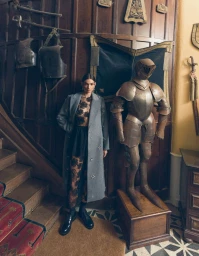 Model wearing a grey embellished coat and sheer floral jumpsuit standing next to a suit of plate armor in a wood-paneled hall.