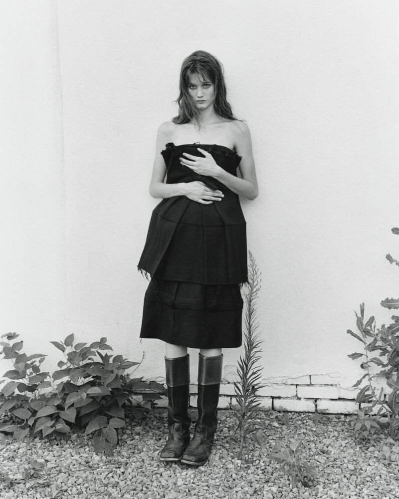 A woman in a dark strapless dress and boots stands outdoors against a white wall with sparse vegetation in a monochrome setting.
