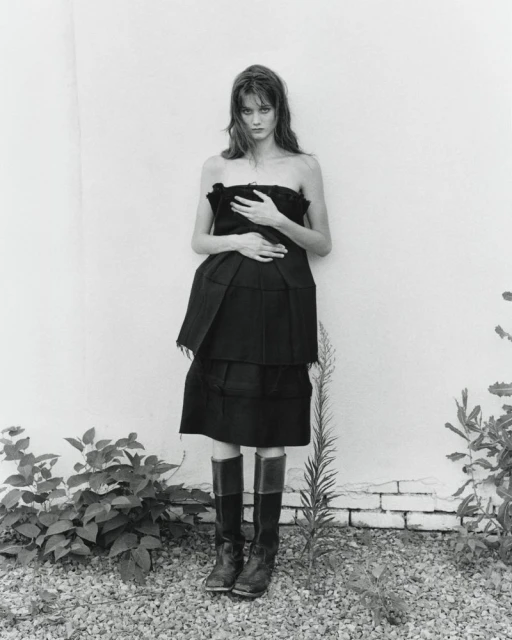 A woman in a dark strapless dress and boots stands outdoors against a white wall with sparse vegetation in a monochrome setting.