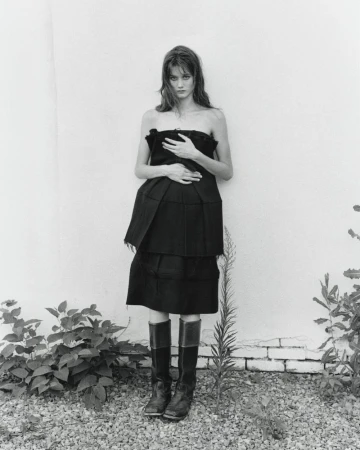 A woman in a dark strapless dress and boots stands outdoors against a white wall with sparse vegetation in a monochrome setting.