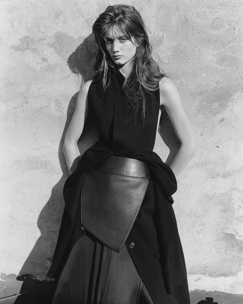 Monochrome portrait of a woman with long hair in black sleeveless top and layered skirt, posing against a textured wall in studio lighting.