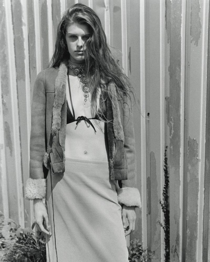 Monochrome fashion editorial portrait of a woman wearing an open shearling jacket and layered necklace against corrugated metal.