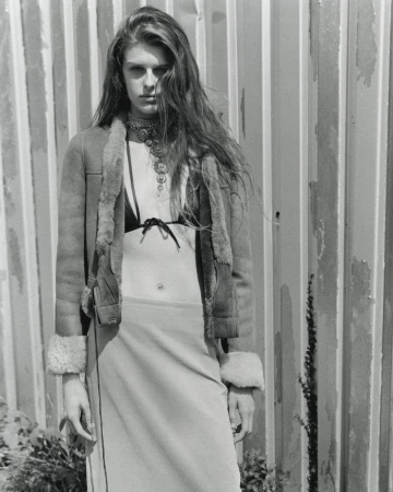 Monochrome fashion editorial portrait of a woman wearing an open shearling jacket and layered necklace against corrugated metal.