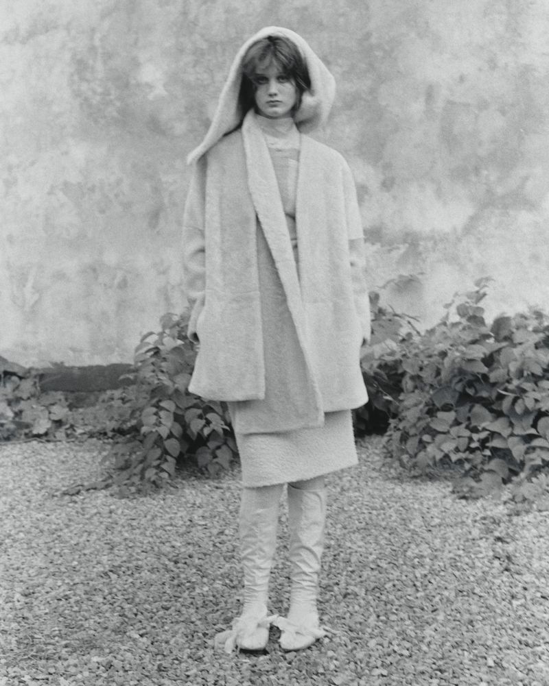 Monochrome portrait of a young woman in hooded coat and boots standing outdoors near greenery, fashion editorial style.