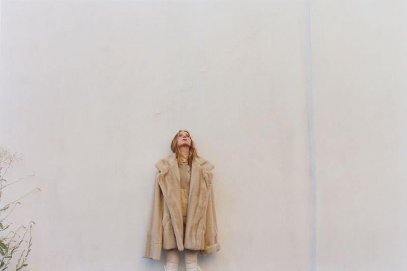 Woman in beige fur coat looking up against a white wall in outdoor fashion photography with minimalist aesthetic, neutral tones, and soft light.