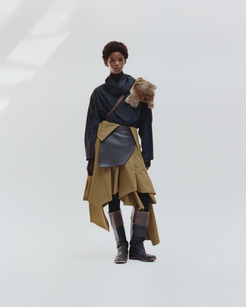 Woman in a long-sleeved top and skirt with boots, posing in a bright studio, with fur accessory draped over her shoulder.