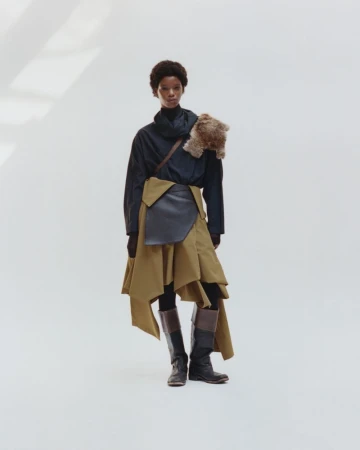 Woman in a long-sleeved top and skirt with boots, posing in a bright studio, with fur accessory draped over her shoulder.