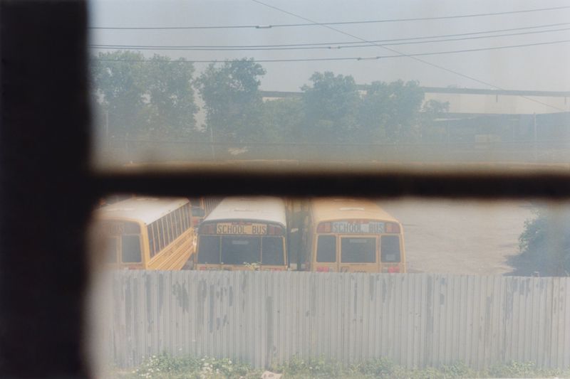 School buses are lined up in a parking lot, viewed through a window, near a metal fence and greenery under a hazy sky.