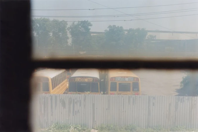 School buses are lined up in a parking lot, viewed through a window, near a metal fence and greenery under a hazy sky.