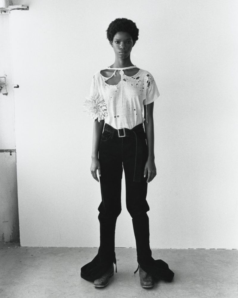 Monochrome full shot of a slim, dark-skinned woman with natural hair posing in a white t-shirt, dark pants, and dress shoes indoors.