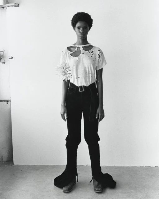 Monochrome full shot of a slim, dark-skinned woman with natural hair posing in a white t-shirt, dark pants, and dress shoes indoors.