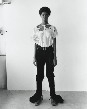 Monochrome full shot of a slim, dark-skinned woman with natural hair posing in a white t-shirt, dark pants, and dress shoes indoors.
