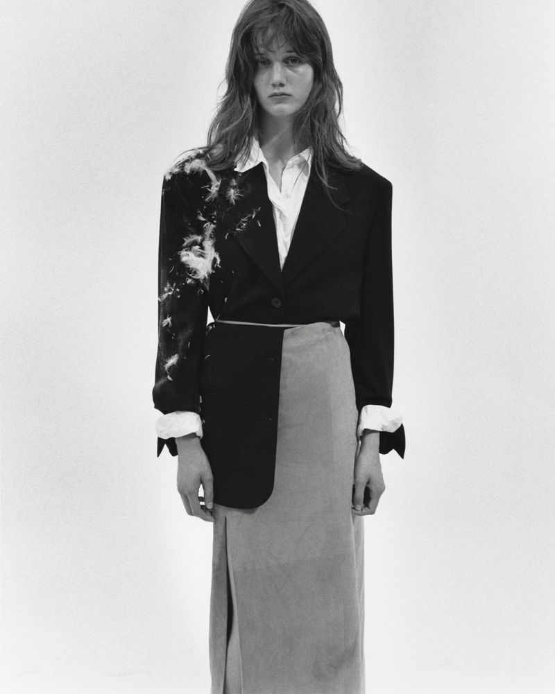 Black and white fashion shot of a long-haired woman wearing a black blazer, white blouse and a long skirt against a white backdrop.