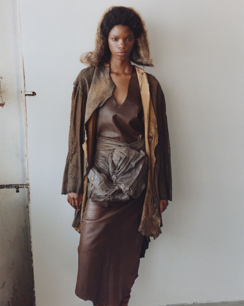A dark-skinned fashion model poses in an earth-toned ensemble in front of an off-white wall; her stare is direct, and her attire is striking.
