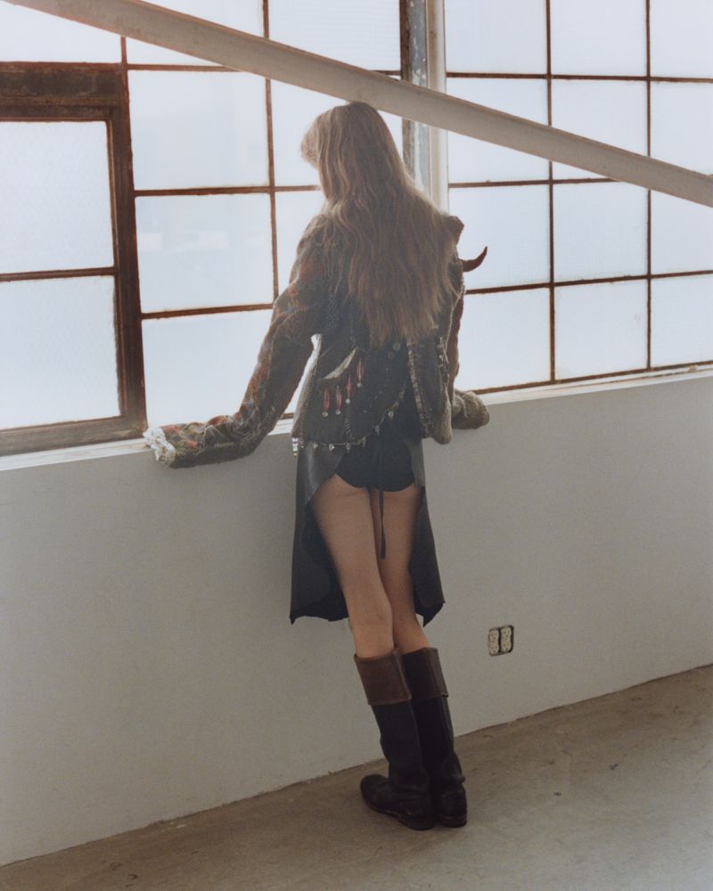 A blonde adult woman stands at a window in a studio wearing a unique vintage-inspired jacket, a short skirt, and tall brown boots.
