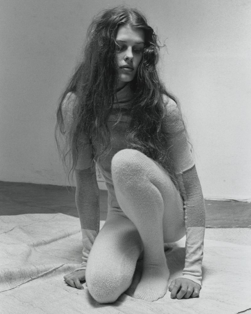 Black and white portrait of a female model crouching on a draped canvas surface in a minimalist studio.