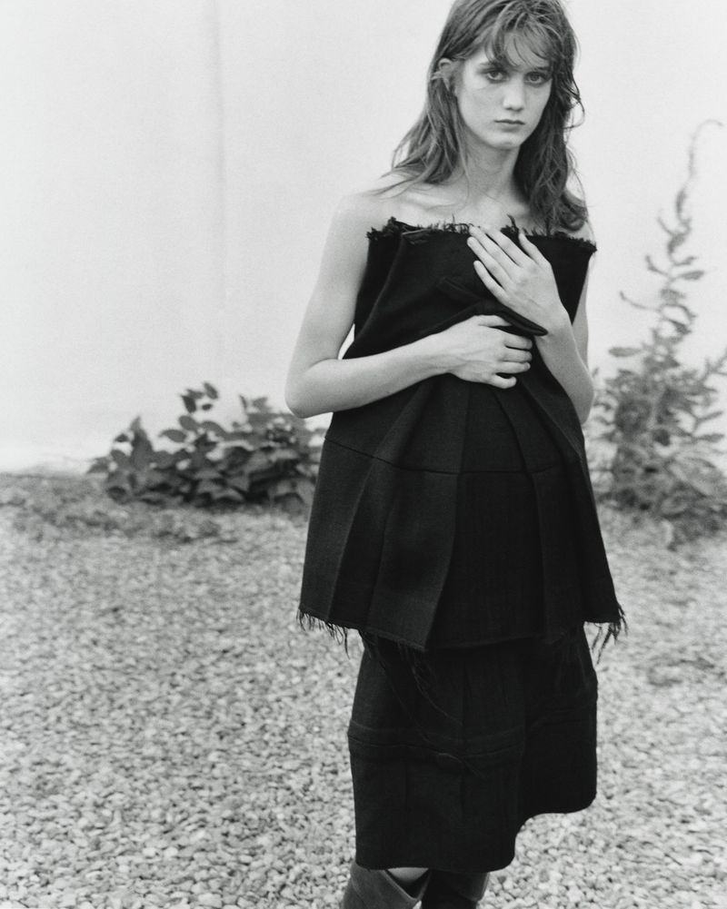 Model in a black strapless pleated top and skirt standing outdoors on gravel against a light wall for The Courtyard project
