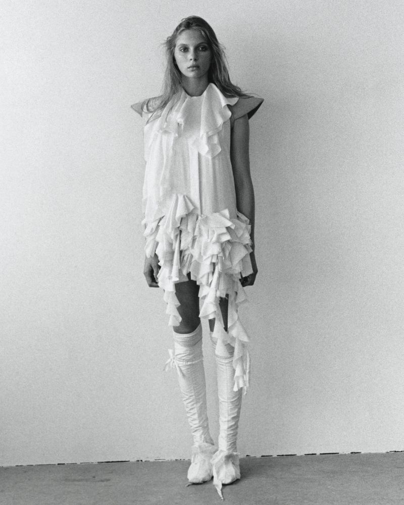 Full-length portrait in black and white of a young woman in a white dress with ruffles, high socks, and neutral expression against white wall.